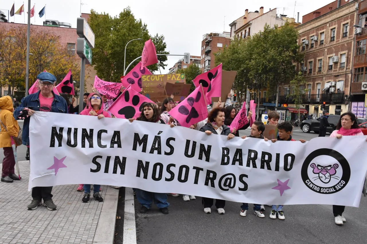 Manifestantes de todas las edades en la Marcha Rosa sosteniendo una gran pancarta blanca con el lema «Nunca más un barrio sin nosotr@s» junto al logo de la Pantera Rosa del Centro Social Seco, rodeados de banderas rosas con huellas negras y carteles reivindicativos.